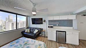Camera d'albergo compatta con un letto, un angolo cottura, una ventola a soffitto, una TV e vista sulla città.
