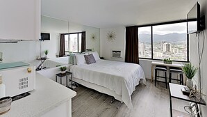 A modern hotel room with a large bed, a desk, and a view of the cityscape through large windows. - Quaint Waikiki Monarch Studio | Pool + Gym (Honolulu)