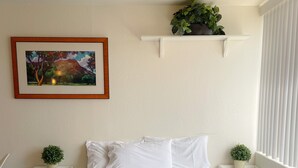 A neatly arranged bedroom with a bed, two bedside tables with potted plants, a framed painting on the wall, and a window with blinds. - Mellow Hawaiian Monarch Studio | Pool + Gym (Honolulu)