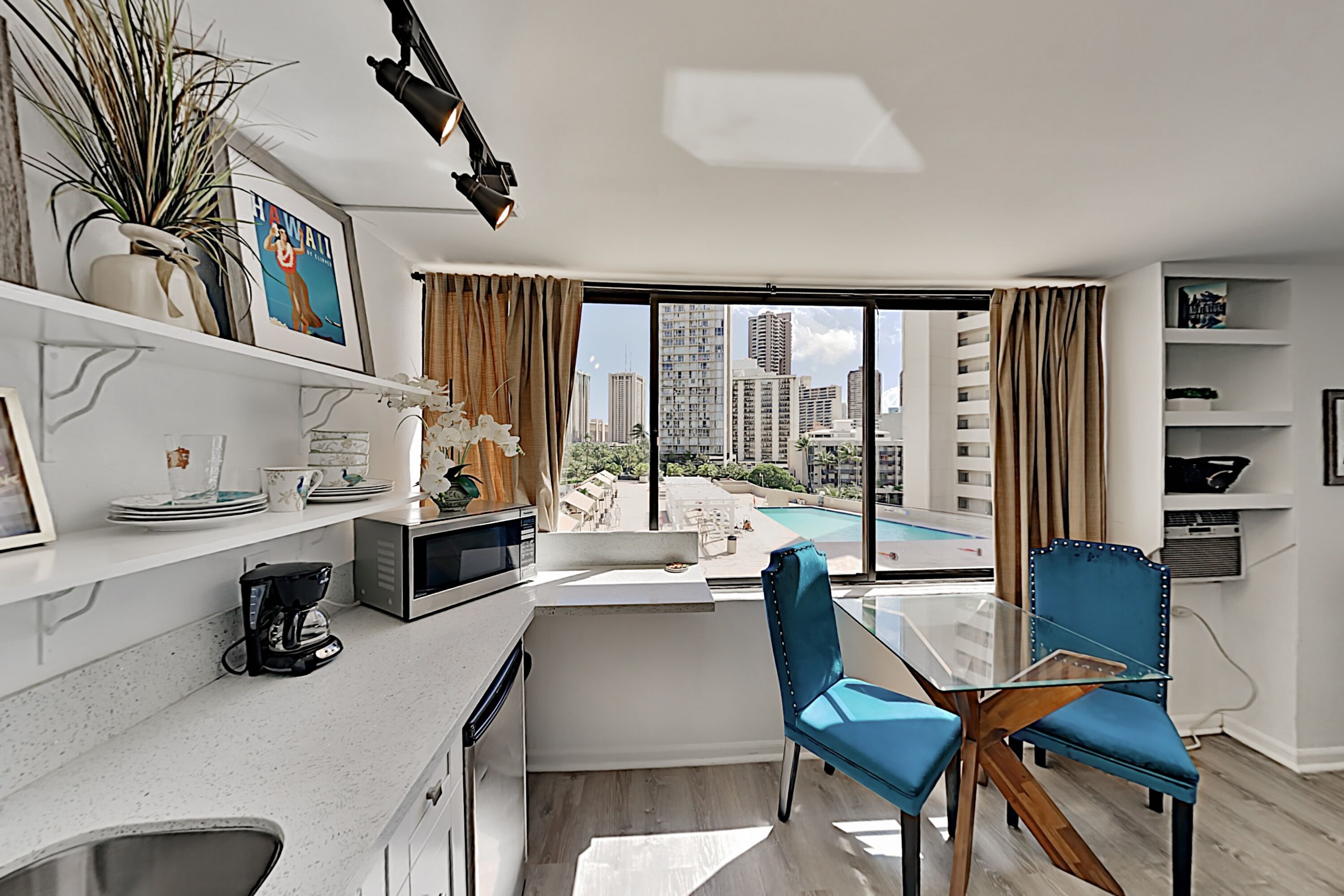 A modern kitchen with a coffee maker, a microwave, and a sink. There is a dining area with a glass table and blue chairs. A large window offers a view of high-rise buildings and a pool.