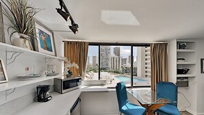 Una cocina moderna con cafetera, microondas y pileta. Hay una zona de comedor con una mesa de vidrio y sillas azules. Un gran ventanal ofrece vistas a edificios altos y una piscina.
