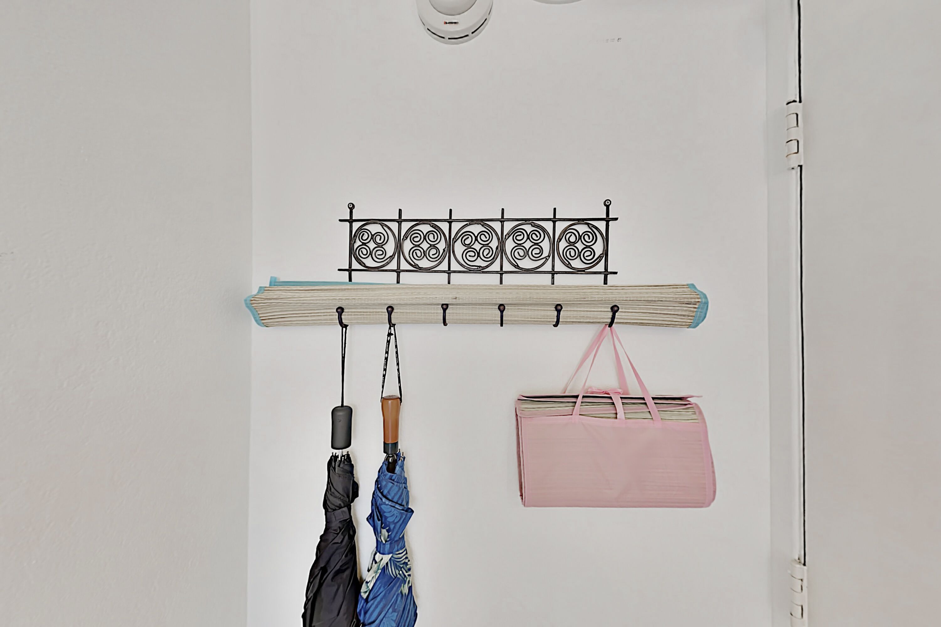A wall-mounted coat rack with a metal decorative piece above it, holding a blue umbrella and a black umbrella. A pink bag hangs on the right side.