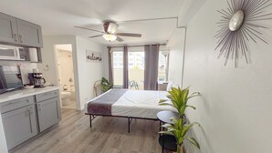 A compact hotel room with a kitchenette, a bed, a ceiling fan, and a view of the outside balcony. - Refreshed! Breezy Kuhio Village Studio | Lanai (Honolulu)