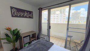 Ein Schlafzimmer mit Bett, kleinem Schreibtisch, Topfpflanze und einer Glastür, die auf einen Balkon mit Blick auf Gebäude führt.