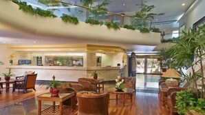 A spacious lobby with wooden flooring, wicker furniture, and potted plants. A reception desk is visible in the background. - Lovely Luana Waikiki Studio | Private Lanai (Honolulu)