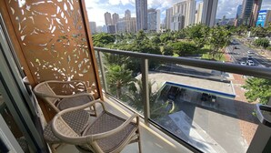 A balcony with wicker chairs, a glass railing, and a city view. - Lovely Luana Waikiki Studio | Private Lanai (Honolulu)