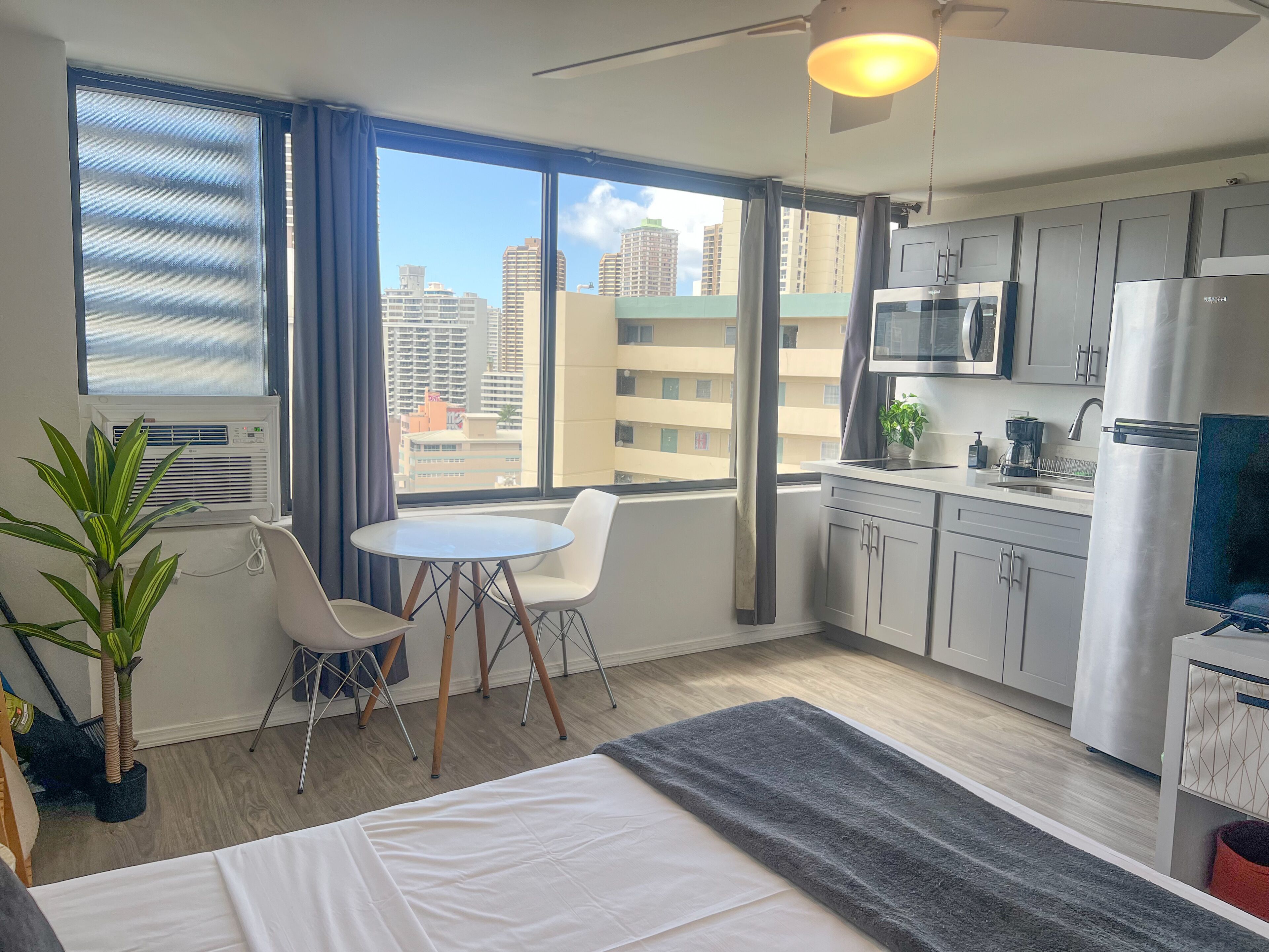 A modern hotel room with a large window, a dining area, a bed, and a kitchenette.