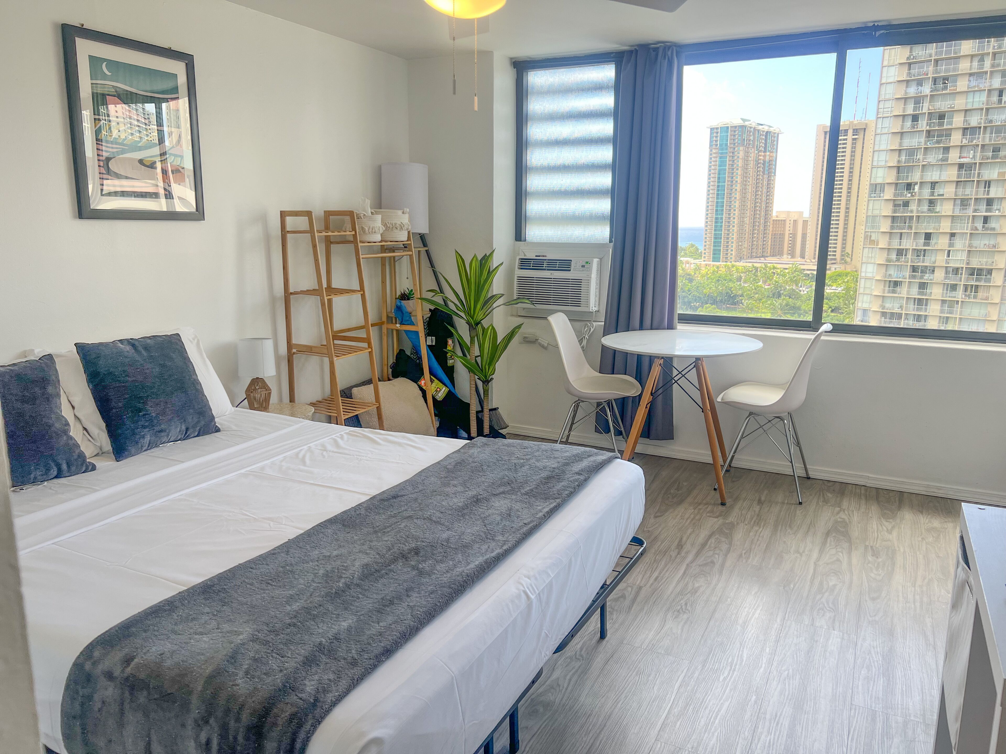 A hotel room with a bed, a small table, two chairs, a plant, a ladder, and a large window with a city view.