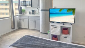 A modern kitchen with grey cabinets, a stainless steel refrigerator, a microwave, and a coffee maker. A flat-screen TV displays a beach scene. - Trendy Hawaiian Monarch Studio | Ocean View | Pool (Honolulu)