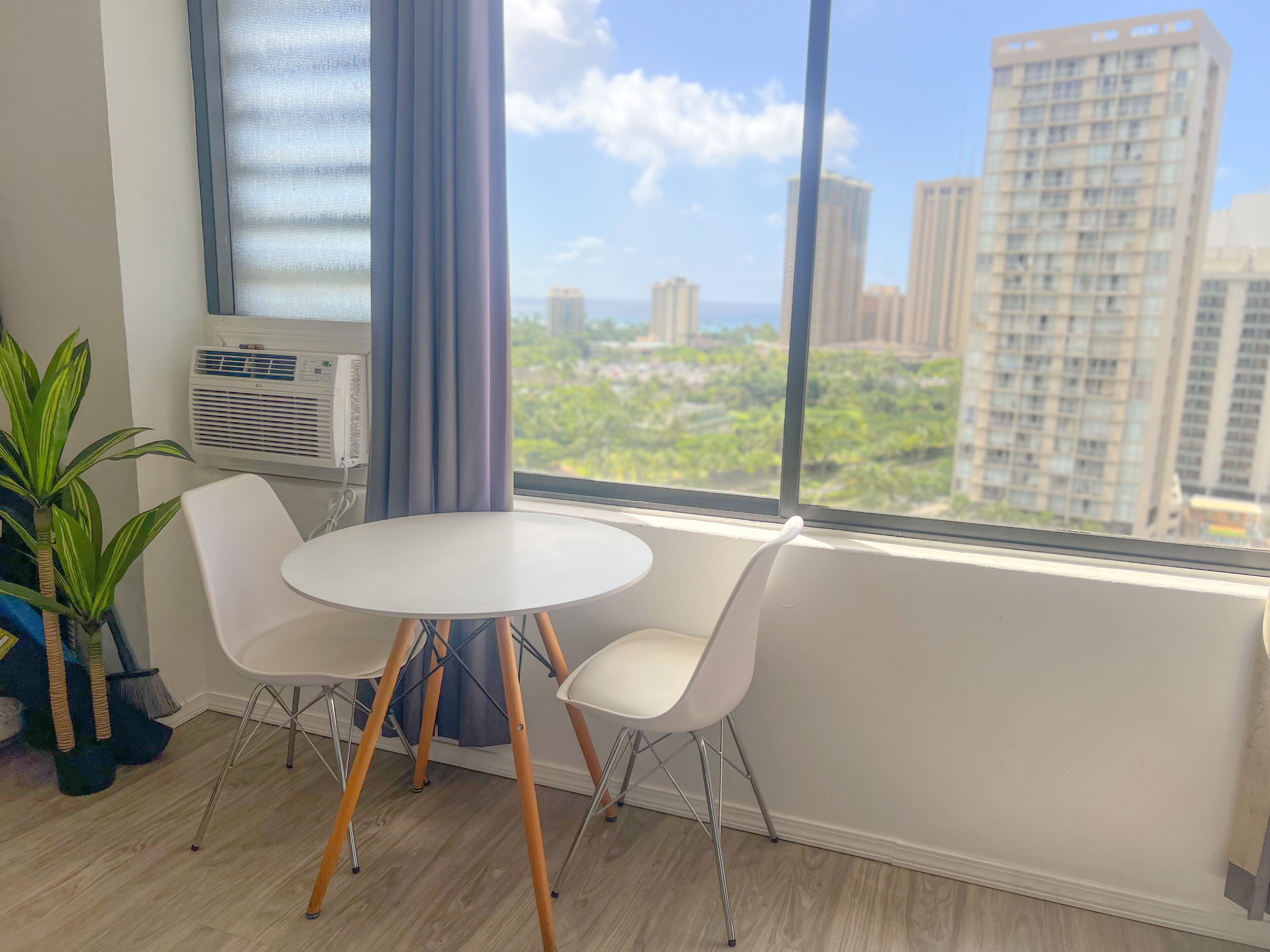 A room with a round white table and two white chairs, a potted plant, and a window with a view of tall buildings and greenery.