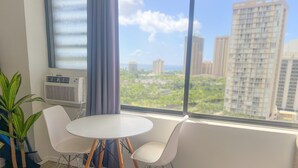 A room with a round white table and two white chairs, a potted plant, and a window with a view of tall buildings and greenery. - Trendy Hawaiian Monarch Studio | Ocean View | Pool (Honolulu)