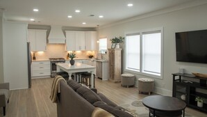 A modern kitchen with white cabinets, a central island, and recessed lighting. There is a dining area with a brown sofa and a small round table. - "Lil House" Cozy 2-bedroom cottage walking distance to beach and downtown. (Bay St. Louis)