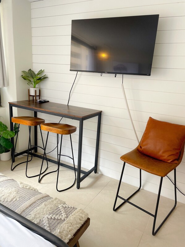 A modern living room with a TV mounted on the wall, a wooden table with two stools, a brown leather chair, and a potted plant.