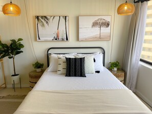 Una cama bien hecha con ropa de cama blanca y almohadas decorativas, dos cuadros con motivos de playa en la pared y una planta en maceta a cada lado de la cama.