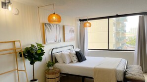 A neatly arranged bedroom with a bed, a bedside table, a hanging plant, and a window with a view of buildings. - Modern Surf Chic Studio at Hawaiian Monarch | Pool (Honolulu)
