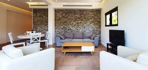 A modern living room with a stone accent wall, a grey sofa, white armchairs, a wooden coffee table, and a flat-screen TV.