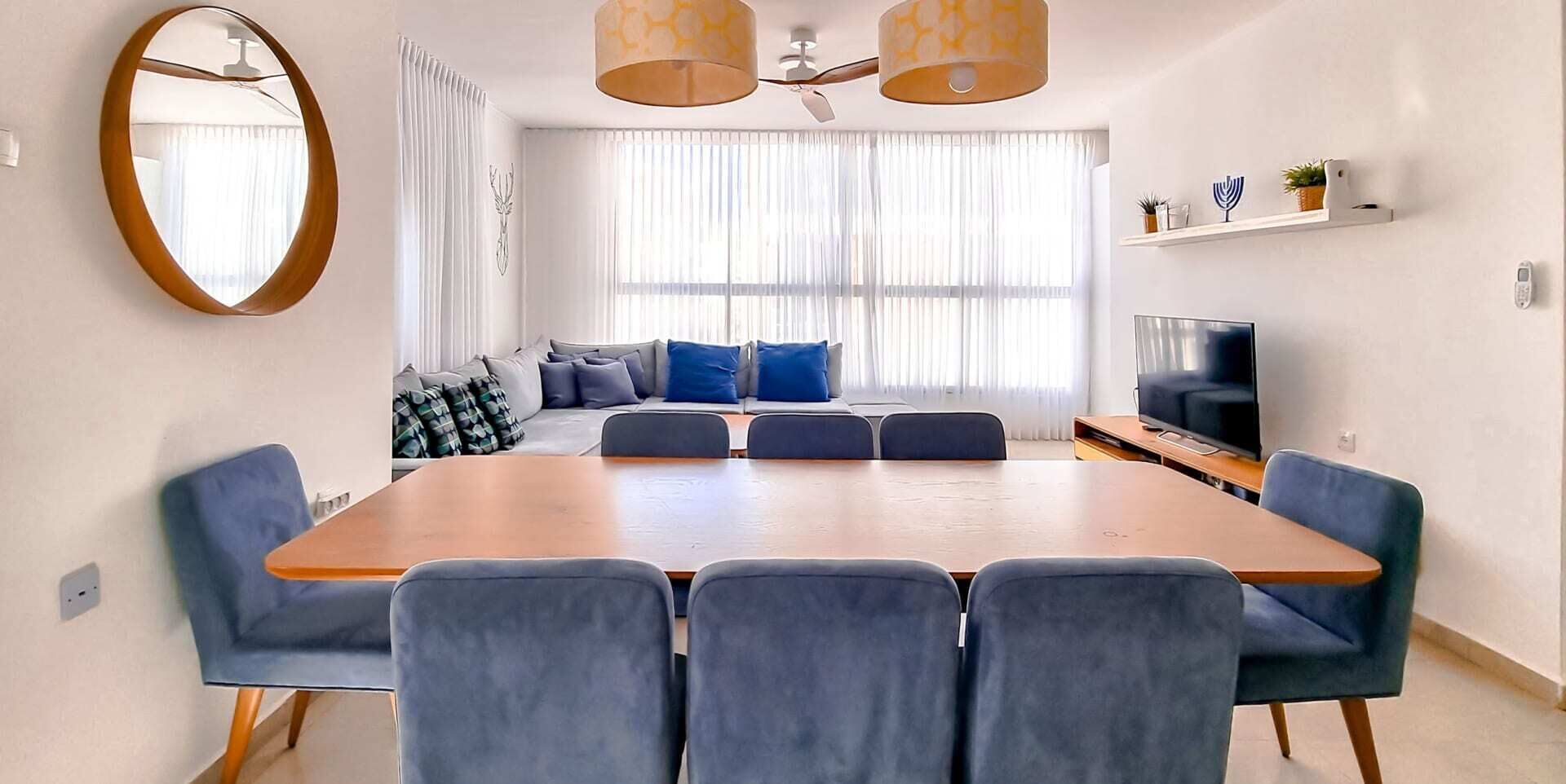 A modern living room with a large wooden dining table, blue upholstered chairs, a sofa with blue cushions, a flat-screen TV, and a ceiling fan.