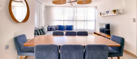 A modern living room with a large wooden dining table, blue upholstered chairs, a sofa with blue cushions, a flat-screen TV, and a ceiling fan.
