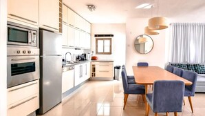 A modern kitchen with built-in appliances, a dining area with a wooden table and blue chairs, and a living area with a sofa and a large window.