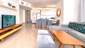 A modern living room with a flat-screen TV, a sofa, a dining area with chairs, and a kitchen in the background.