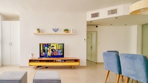 A modern living room with a flat-screen TV, a sofa, two ottomans, and a dining area with blue chairs.
