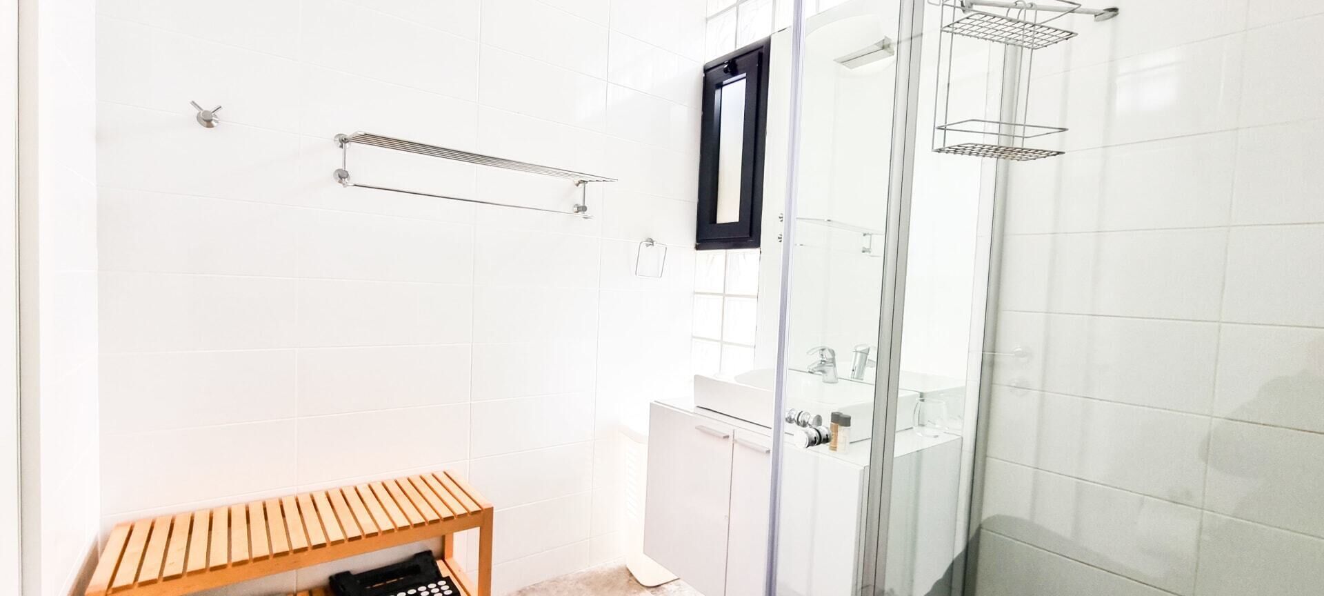 A modern bathroom with white tiles, a glass shower enclosure, a wooden bench, and a towel rack.