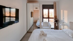 Ein Hotelzimmer mit großem Flachbildfernseher an der Wand, einem Bett mit weißer Bettwäsche, einem Sessel, einem Schreibtisch mit Stuhl und einem Fenster mit Blick auf Gebäude.