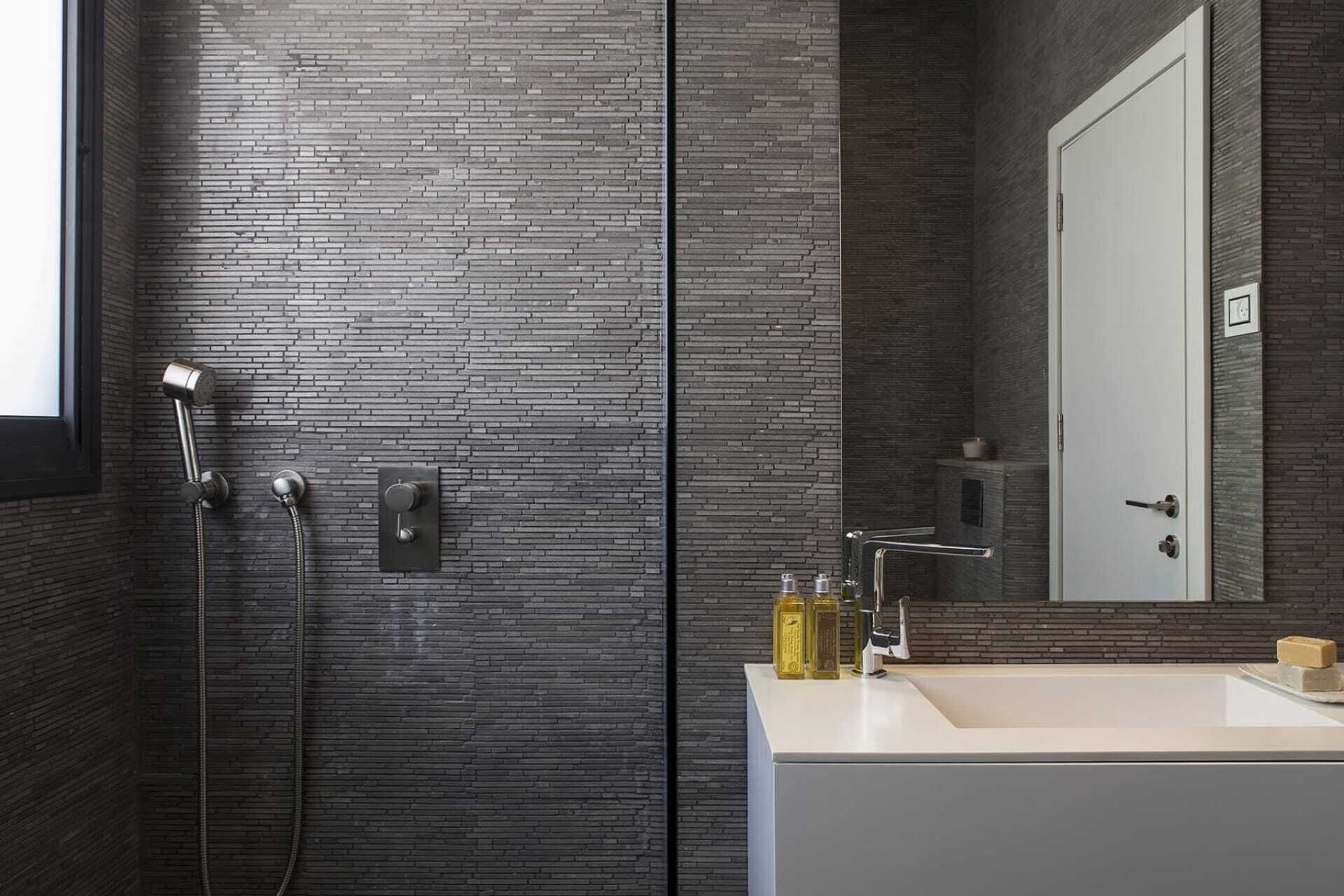 Un baño moderno con pared de ladrillo, ducha con cabezal de mano, lavamanos con espejo y artículos de aseo en la encimera.