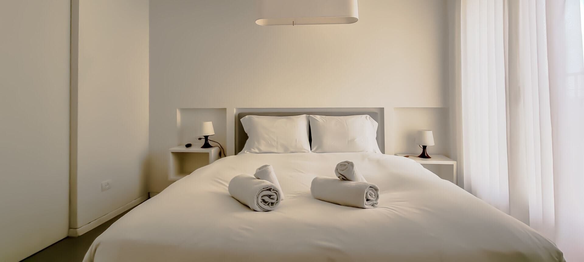 A neatly made bed with rolled towels, a bedside table with a lamp, and a large window with curtains.
