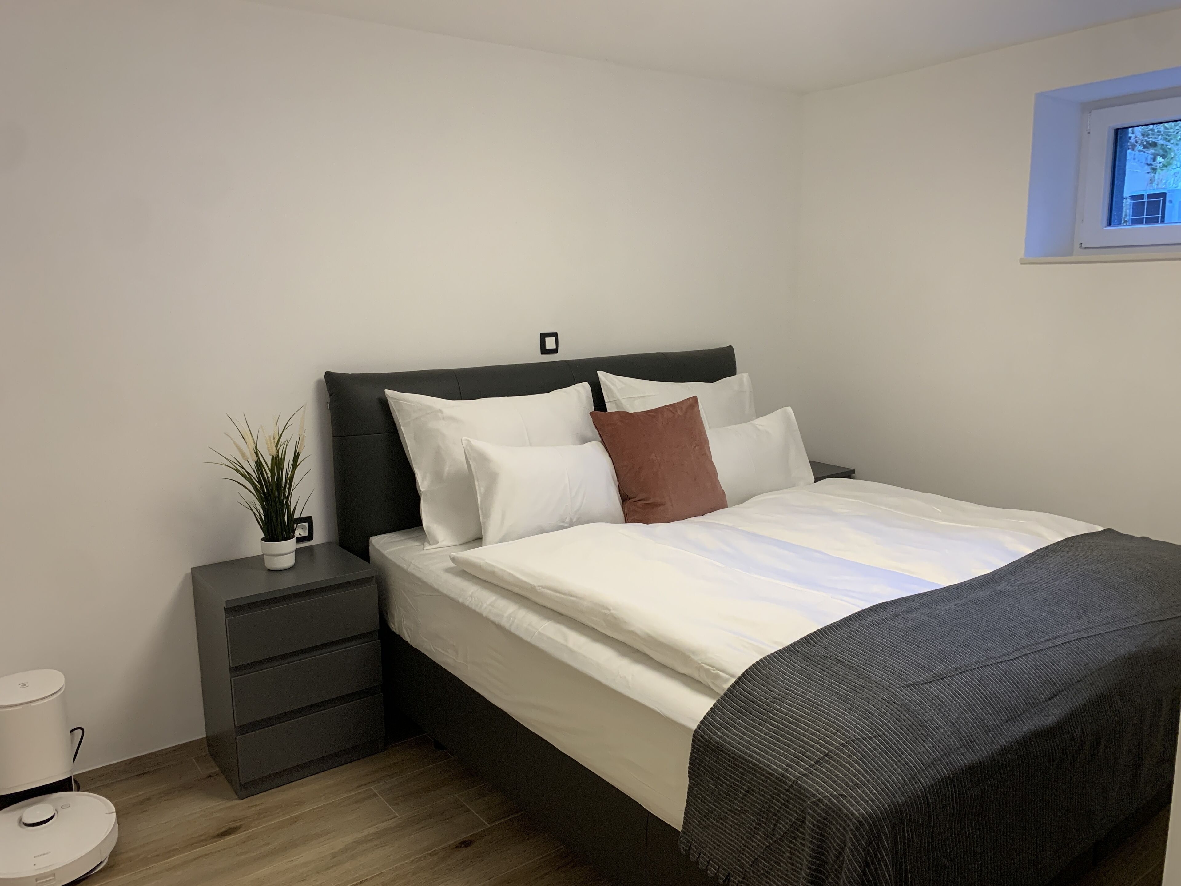 A neatly made bed with white and brown pillows, a dark headboard, and a grey blanket. There is a small bedside table with a plant and a charging device.