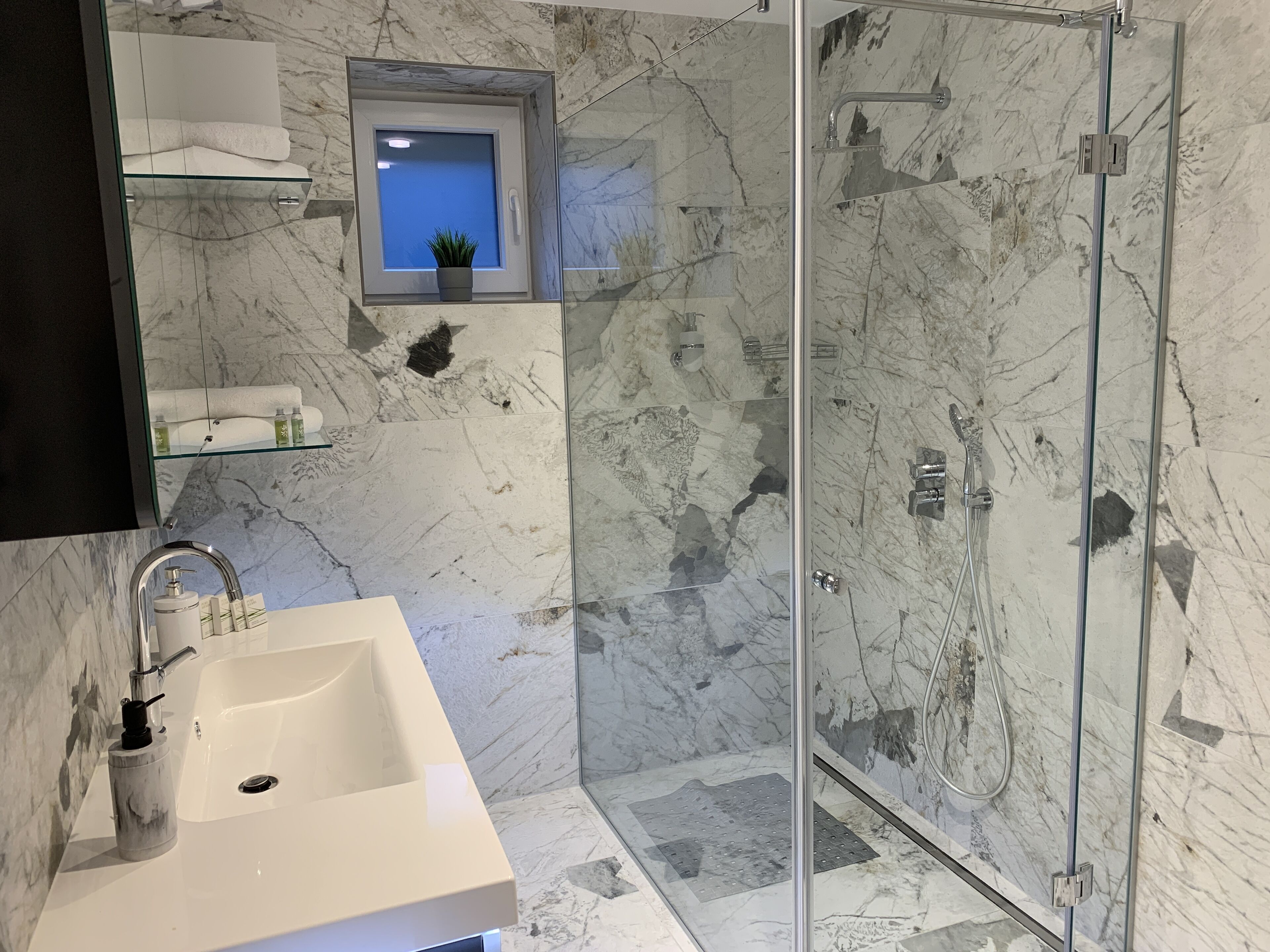 Ein Badezimmer mit einer Duschkabine, einem Waschbecken mit Marmorplatte und einem kleinen Fenster mit Pflanze.