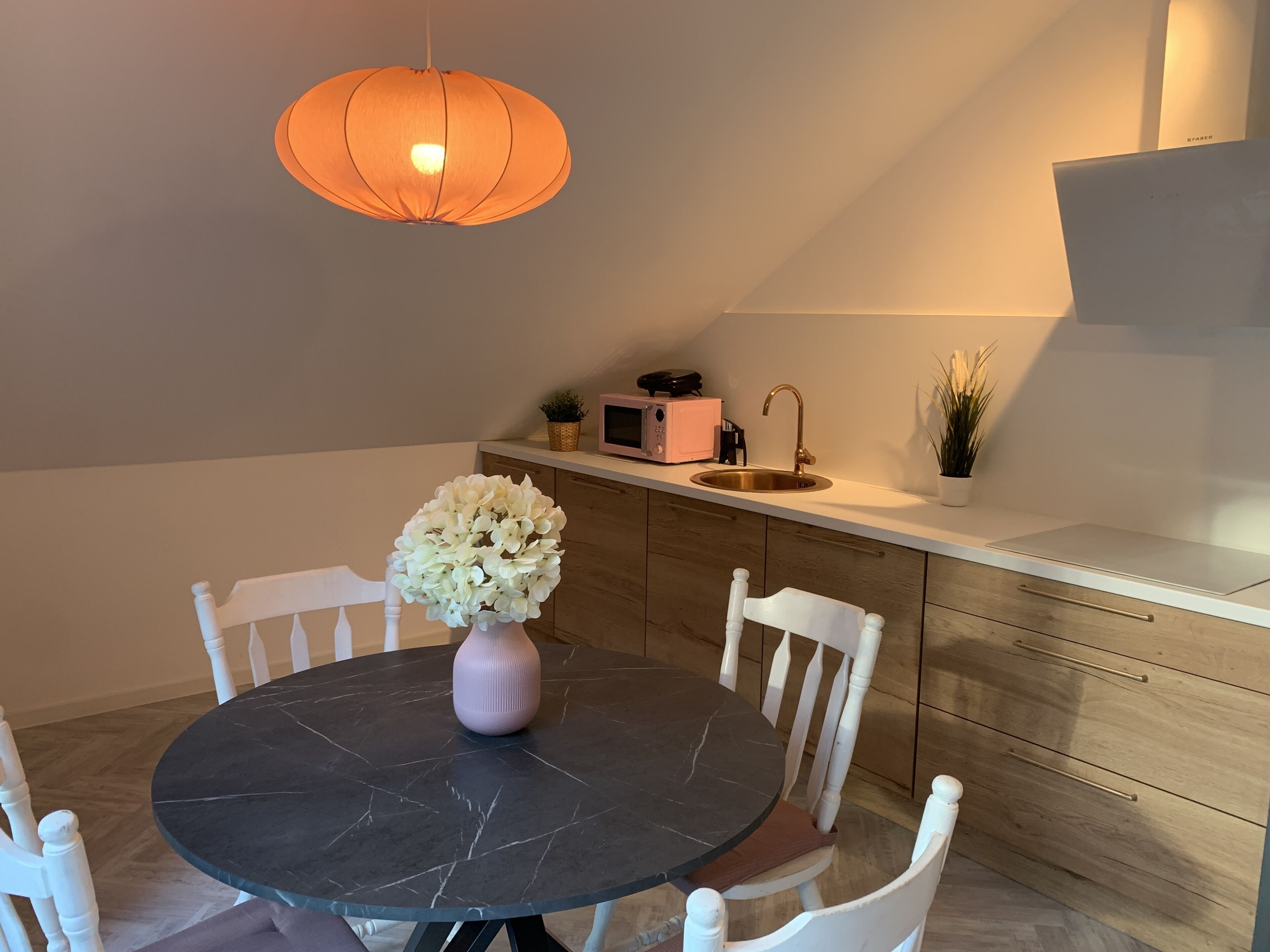 Een eetruimte met een ronde tafel, witte stoelen, een bloemenvaas, een hanglamp en een keuken op de achtergrond.