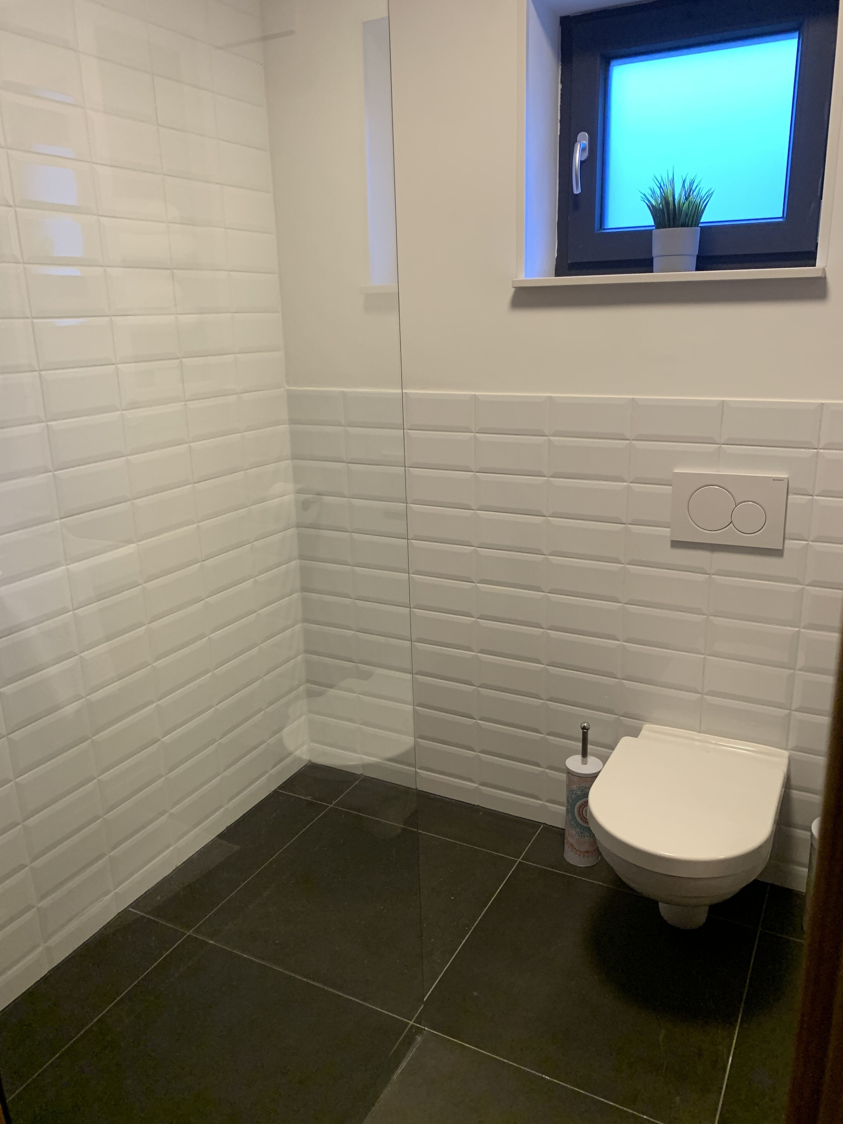 Une salle de bain avec des murs recouverts de carreaux blancs, des toilettes blanches et une petite fenêtre avec une plante.