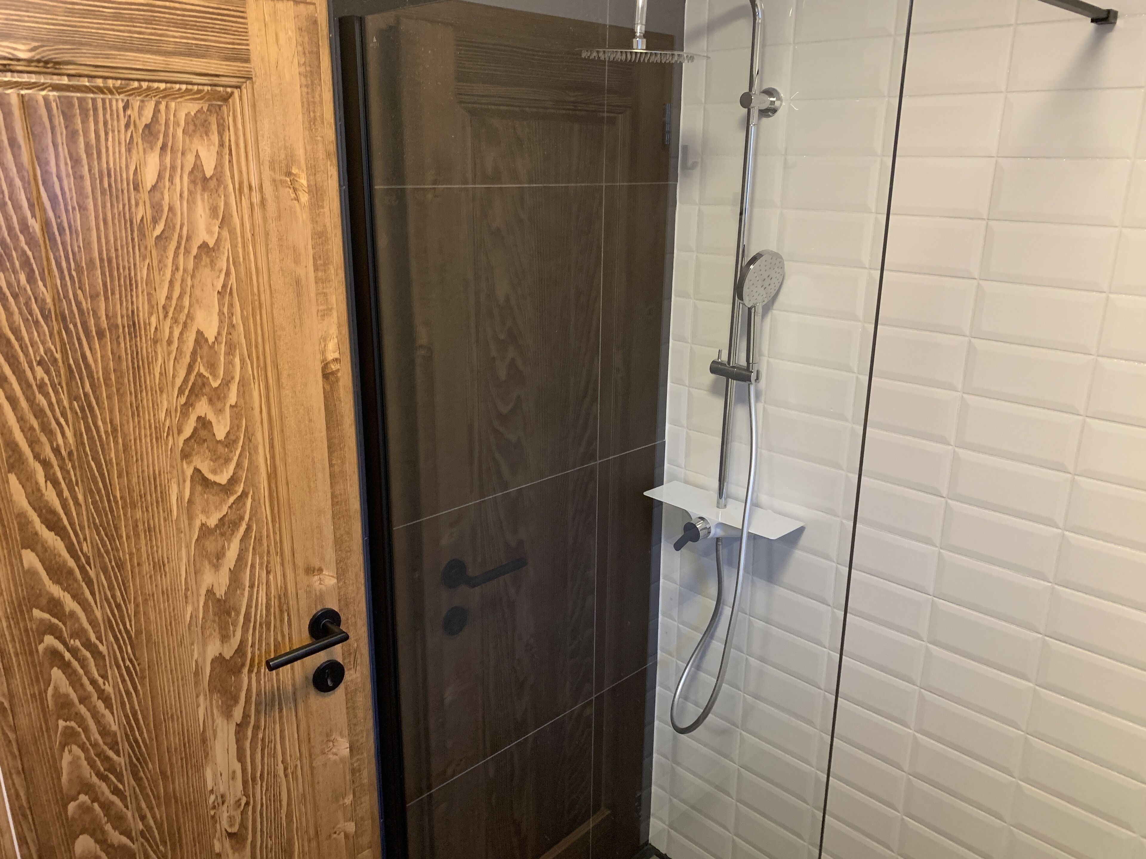 Une porte en bois donne accès à un espace douche dont les murs sont recouverts de carreaux blancs et qui est doté d’une porte en verre.
