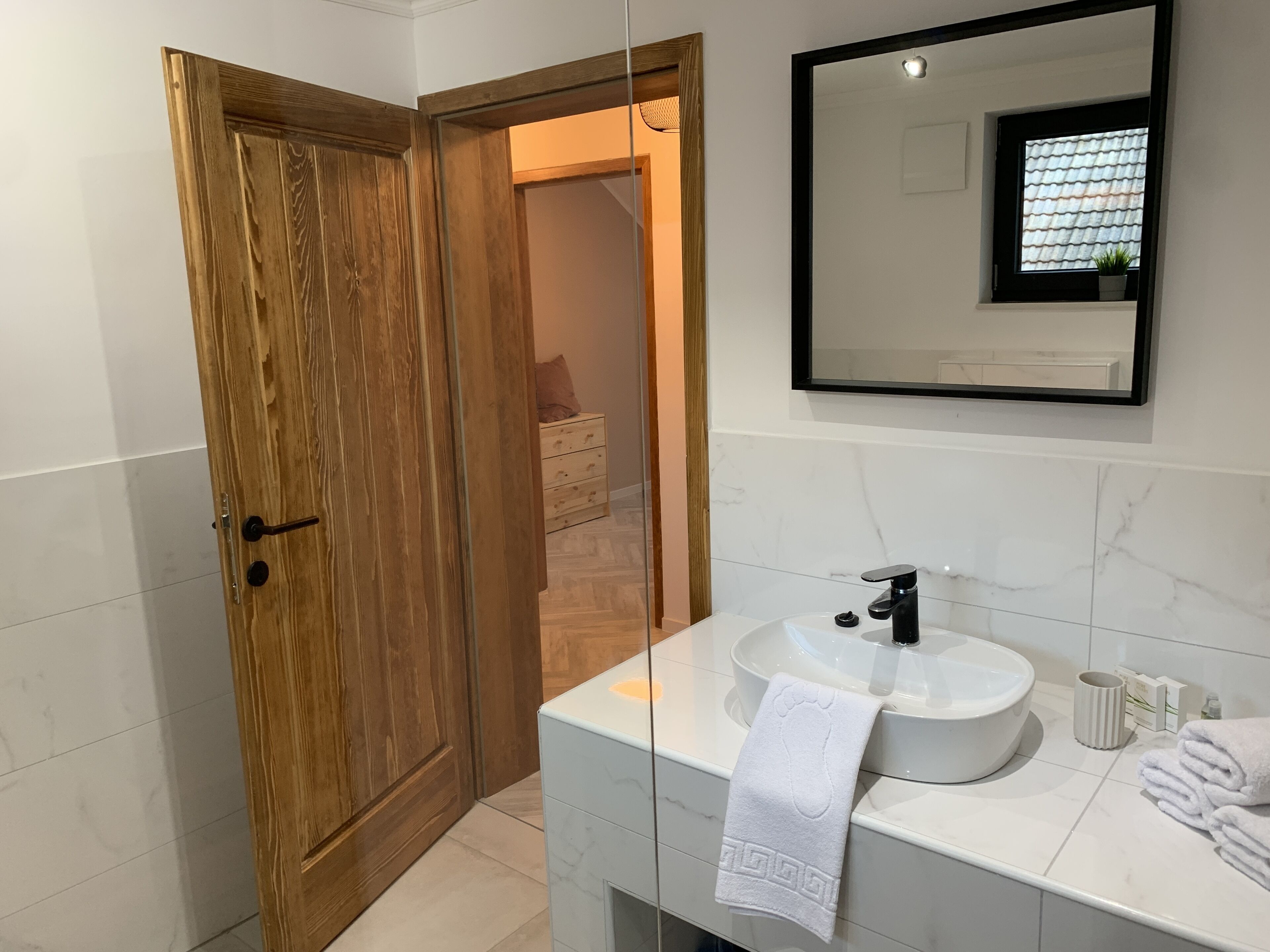 Een moderne badkamer met een houten deur, een witte wastafel, een spiegel en een handdoekrek.