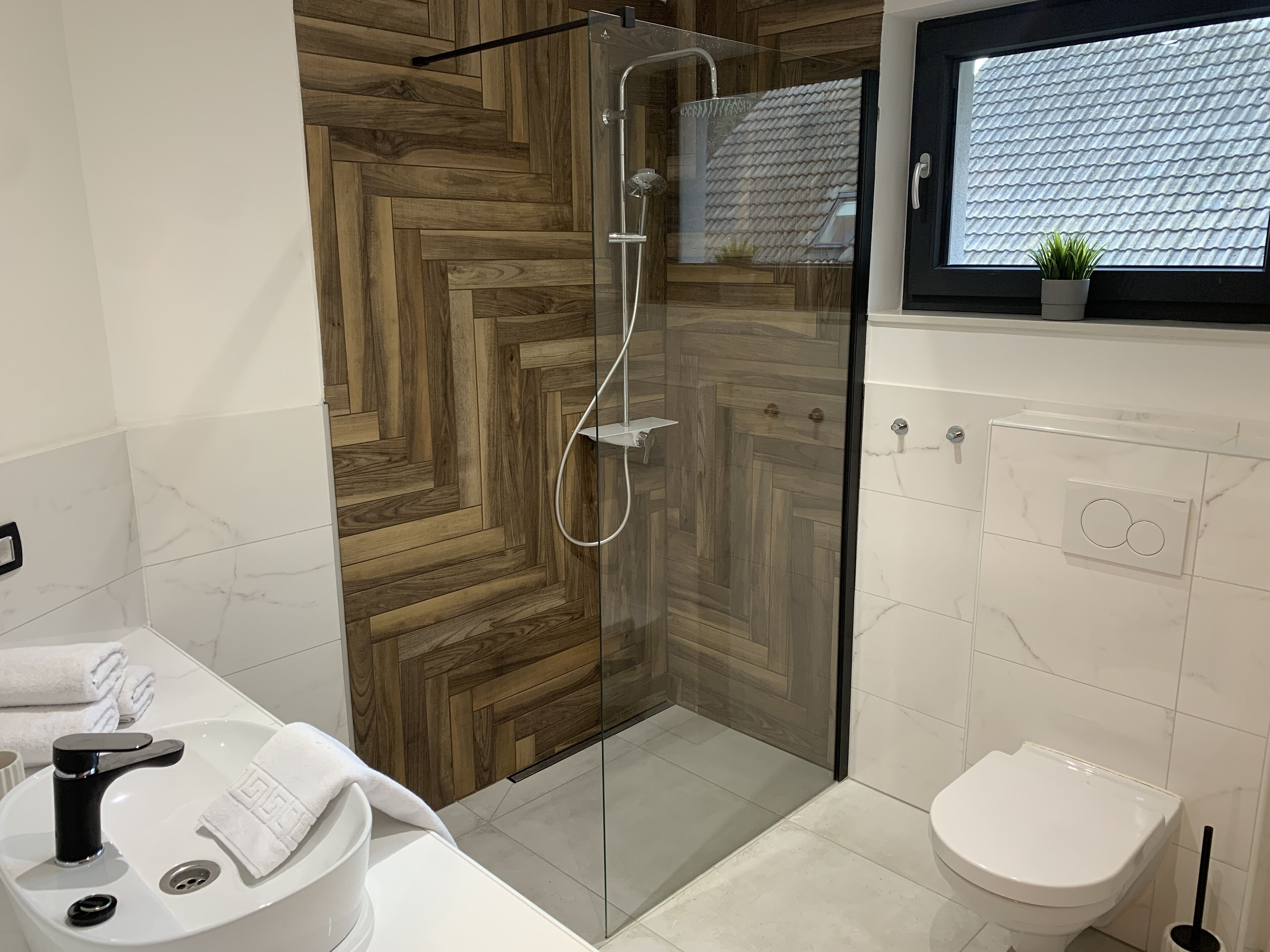 Une salle de bain moderne avec une douche à parois de verre, un lavabo blanc avec un robinet noir, des toilettes et une fenêtre donnant sur un toit recouvert de tuiles.