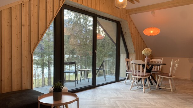 A cozy room with a large sliding glass door leading to an outdoor deck, wooden walls, a round table with chairs, and a small round table with a plant. - Pine Tree Holiday House (Kranjska Gora)