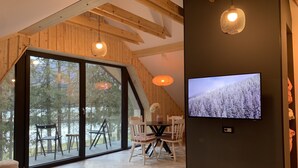 A modern interior with a large sliding glass door leading to a deck, wooden beams on the ceiling, a dining area with white chairs, and a flat-screen TV mounted on a dark wall. - Pine Tree Holiday House (Kranjska Gora)