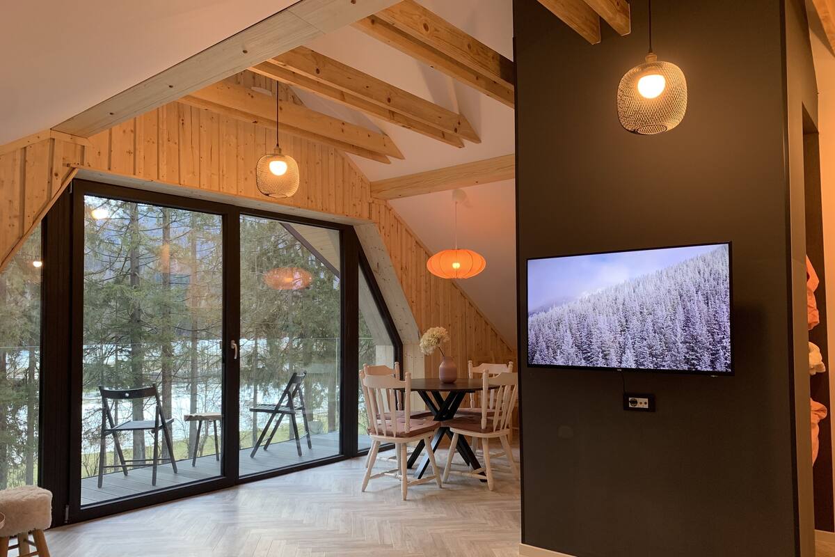 Un interior moderno con un amplio ventanal corredera que da a una terraza, vigas de madera en el techo, una zona de comedor con sillas blancas y un televisor de pantalla plana montado en una pared oscura.