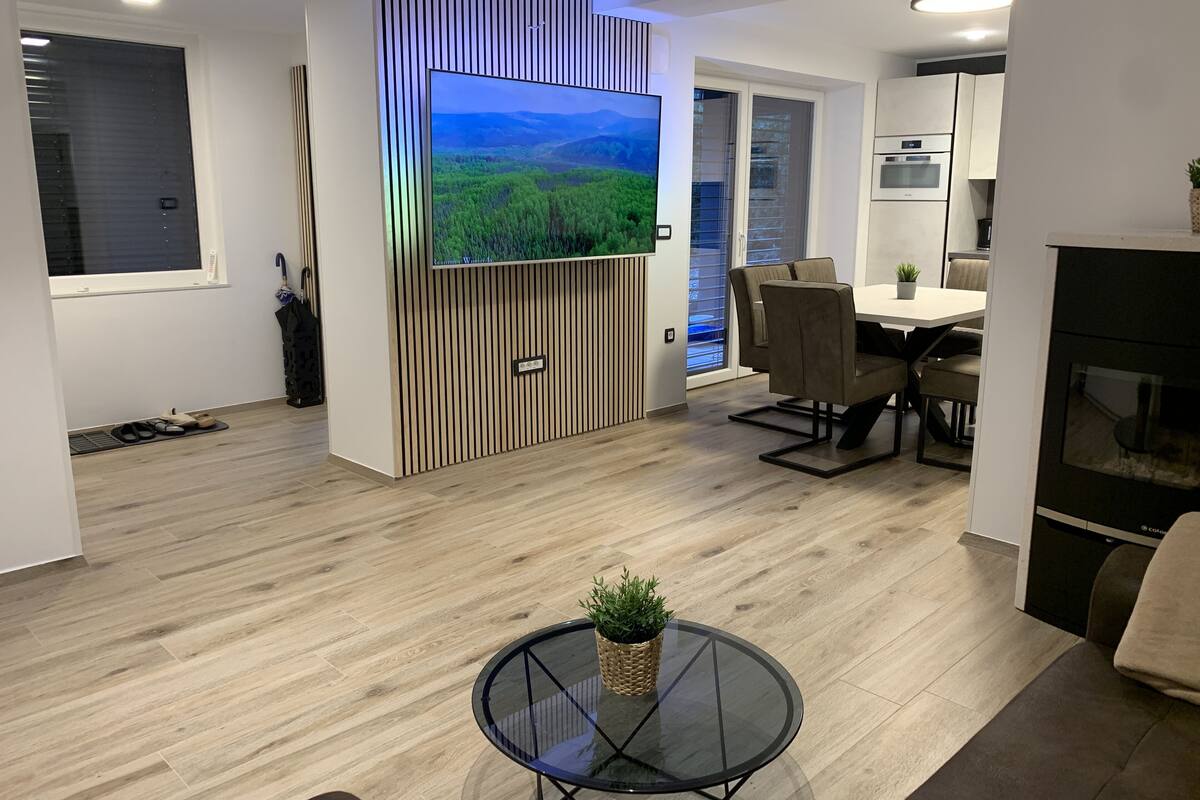 Una sala de estar moderna con un televisor de pantalla plana montado en un panel de madera, un comedor con mesa y sillas, y una cocina con electrodomésticos integrados.