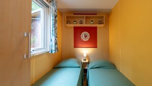 A small, narrow room with two beds, a window with curtains, a shelf with decorative items, and a wall clock. - Estrellus, mobile home in the vineyard (Hyères)