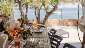 Un espace de restauration en plein air avec une table dressée pour le petit-déjeuner, donnant sur une étendue d’eau où l’on aperçoit un bateau au loin.