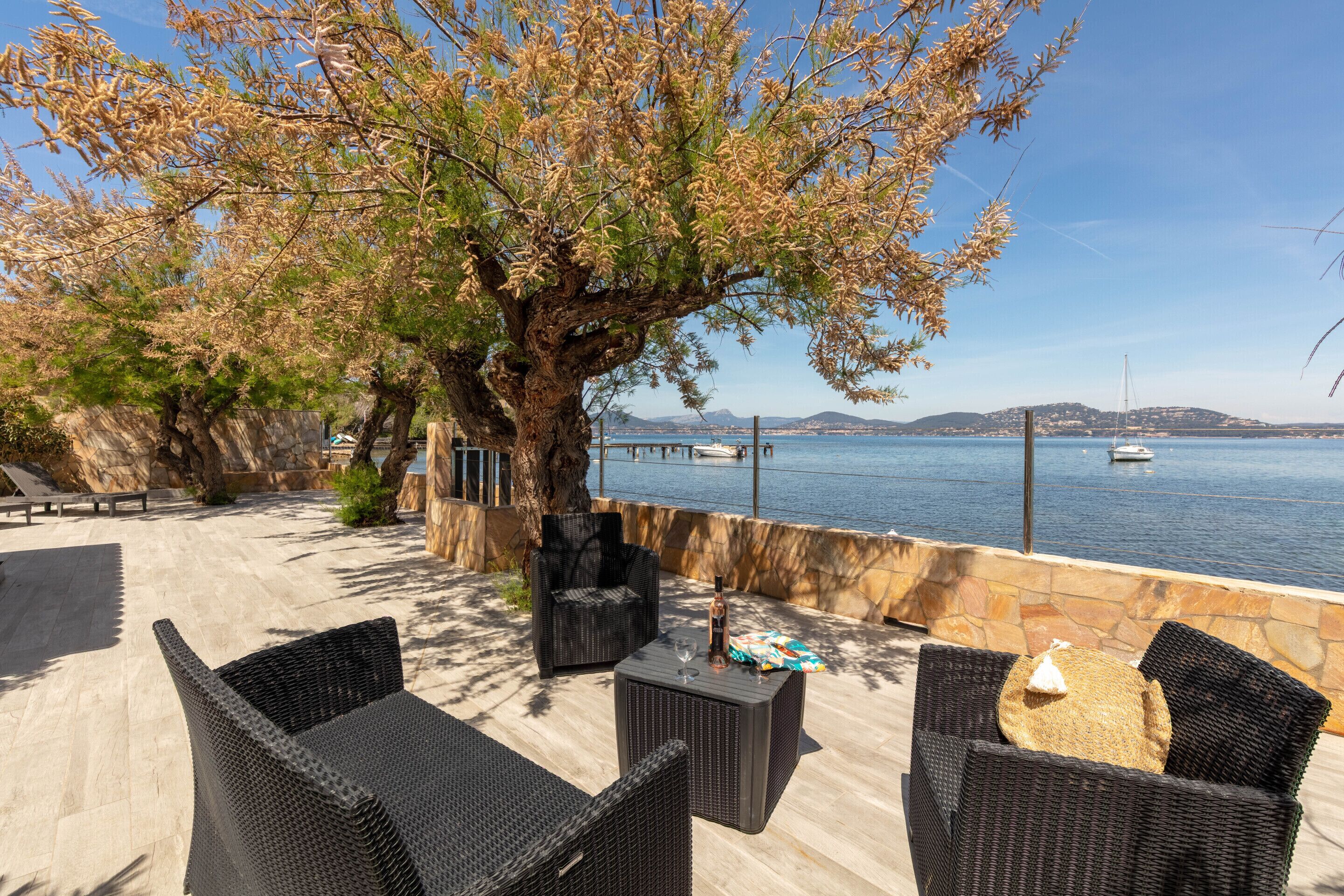 An outdoor seating area with wicker furniture, a stone wall, and a view of the sea with boats.