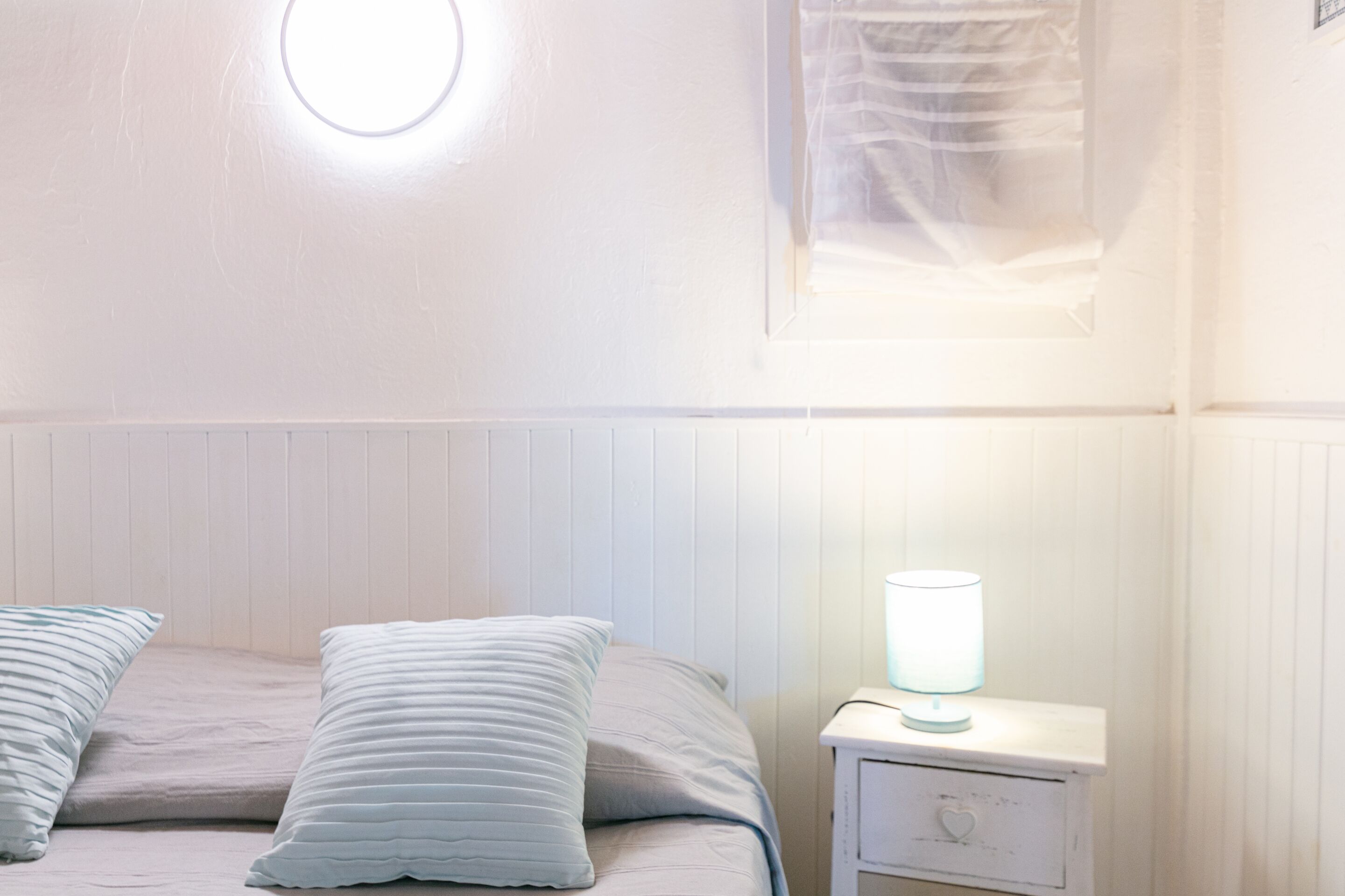 A bedroom with a bed, two pillows, a bedside table with a lamp, and a window with blinds.
