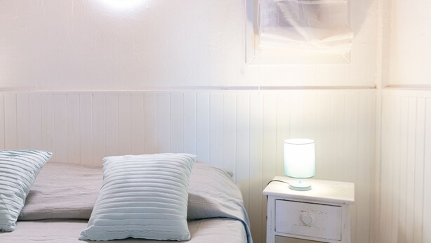Ein Schlafzimmer mit einem Bett, zwei Kissen, einem Nachttisch mit Lampe und einem Fenster mit Jalousien.
