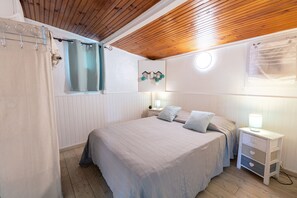 A bedroom with a wooden ceiling, a bed with a light-colored bedspread, two bedside tables with lamps, and a window with a curtain.