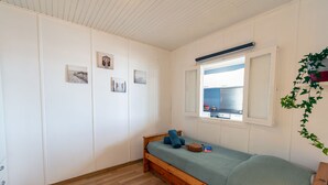 Une petite chambre avec un lit simple, un cadre en bois, une fenêtre donnant sur un immeuble et un mur orné de trois tableaux.