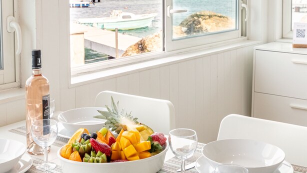 Una mesa de comedor con frutero, botella de vino y copas, con vista a la playa.