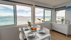 Un coin repas avec une table dressée pour deux personnes, de grandes fenêtres donnant sur la mer et une horloge au mur.