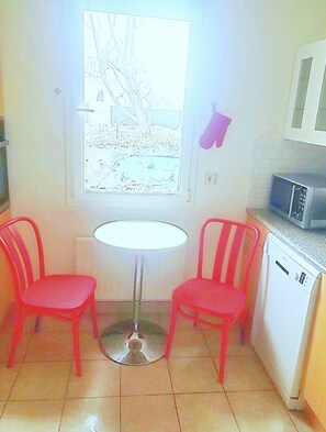 Une petite cuisine avec deux chaises rouges, une table blanche, une fenêtre donnant sur des arbres, un four à micro-ondes et un lave-vaisselle.